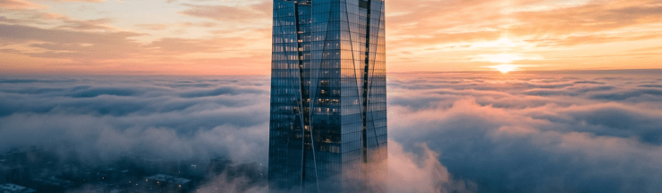 Tall glass skyscraper emerging above dense fog at sunrise